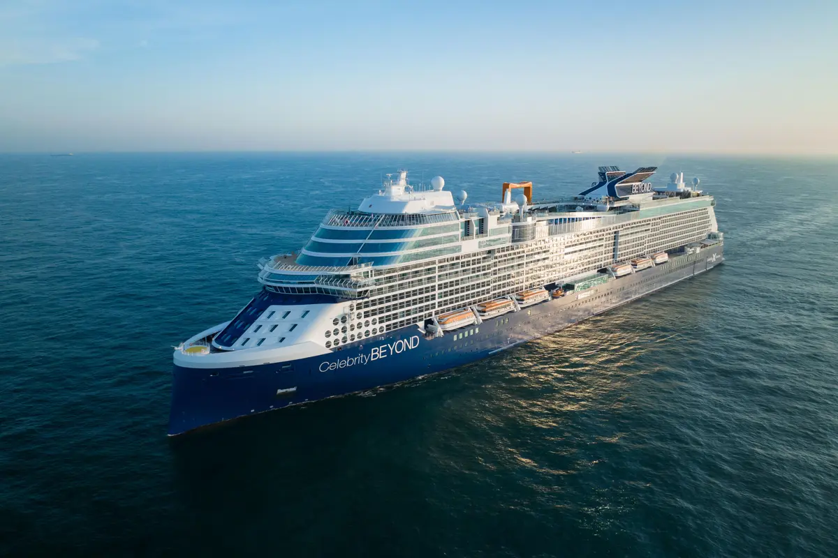 Ein großes, blau-weißes Kreuzfahrtschiff namens Celebrity Beyond fährt auf offenem, blauem Meer unter klarem Himmel. Die Morgensonne spiegelt sich auf dem Wasser und den oberen Decks wider. Das Schiff ist leicht nach links geneigt und wird von oben fotografiert.