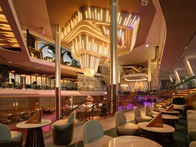 Das moderne, mehrstöckige Atrium der Celebrity Beyond. Eine große, beleuchtete Deckeninstallation dominiert den Raum über eleganten Lounge-Bereichen.