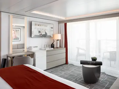 Eine helle und moderne Balkonkabine mit einem großen Bett im Vordergrund, einem Schreibtisch, einer Kommode mit Fernseher und einem Balkon mit Vorhängen im Hintergrund. Ein Yogamatte und eine Pflanze stehen auf einem Beistelltisch.