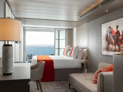 Moderne Kabine auf der Celebrity Beyond. Sie zeigt ein Bett, Schreibtisch, Lounge und Balkon mit Meerblick. Helle Einrichtung mit Kunstwerk an der Wand.