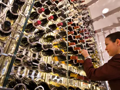 Eine Frau wählt Wein aus einem hohen, modern gestalteten Regal. Hunderte Flaschen in beleuchteten Metallhaltern sind sichtbar und schaffen eine elegante Atmosphäre.