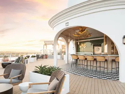Abendstimmung auf der Celebrity Beyond: Eine weiße Outdoor-Bar mit Bögen, Rattan-Hockern und gemustertem Boden, daneben moderne Lounge-Möbel und Pflanzen unter warmem Himmel.