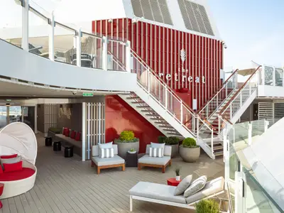 Sonniges Außendeck der Celebrity Beyond. Rote Fassade mit 'The Retreat'-Schriftzug, weiße Treppe, diverse Loungemöbel und Pflanzen.