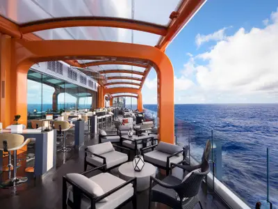 Modernes Außendeck der Celebrity Beyond. Orangefarbene Träger, Glasdach, Loungemöbel, Bar und Blick auf blaues Meer sowie Himmel mit Wolken.