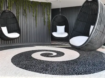 Drei schwarze Korbsessel mit weißen Kissen in einer Lounge. Der Boden zeigt ein schwarz-weißes Spiralmuster aus Kieselsteinen. Dunkle Lamellenwand und hängende Pflanzen im Hintergrund.