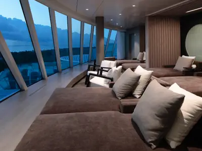 Eine geräumige Lounge auf der Celebrity Beyond mit mehreren Reihen bequemer, brauner Liegen und Kissen. Große, schräge Panoramafenster bieten einen weiten Blick auf das Meer und die Küste in der Dämmerung. Zwei moderne Sessel sind ebenfalls zu sehen.