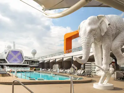 Pooldeck Celebrity Beyond mit Pool, Liegestühlen und weißer Elefantenstatue auf menschlicher Figur. Moderne Schiffselemente, orange Struktur und Bar im Hintergrund.