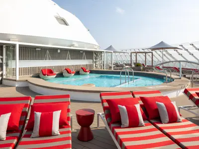 Das sonnige Pooldeck der Celebrity Beyond mit geschwungenem Pool, rot-beige gestreiften Liegen, roten Kissen und weißen Hängesesseln. Im Hintergrund moderne Schiffarchitektur.