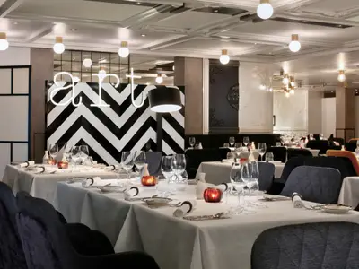 Modernes Restaurant auf der Celebrity Beyond. Gedeckte Tische, dunkle Stühle, chevron-gemusterte Wand mit 'art'-Schriftzug. Helle Deckenleuchten.