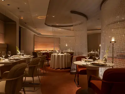Ein elegantes Restaurant mit vielen runden Tischen, die mit weissen Tischdecken und Geschirr gedeckt sind. Die Beleuchtung ist warm und gedimmt. Grosse, durchsichtige Paravents schaffen eine intime Atmosphäre. Keine Personen sind sichtbar.