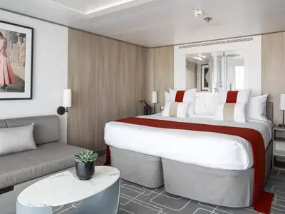 Helle, modern gestaltete Kabine auf der Celebrity Beyond mit einem großen Doppelbett, rotem Überwurf und grauem Sofa. Ein Couchtisch mit Pflanze und ein gerahmtes Bild schmücken den Raum.