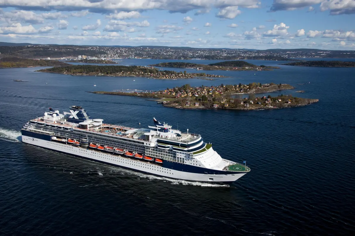 Luftaufnahme des Kreuzfahrtschiffs Celebrity Constellation, das auf ruhigem, blauem Wasser fährt. Im Hintergrund sind grüne Inseln und eine Stadt an der Küste zu sehen.