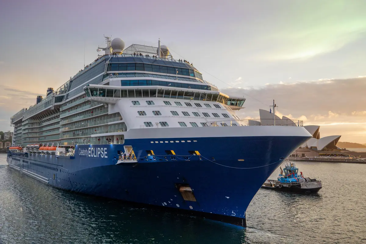 Ein grosses blau-weisses Kreuzfahrtschiff, die Celebrity Eclipse, liegt im Hafen. Rechts ist ein kleiner Schlepper zu sehen, und im Hintergrund die markante Architektur der Oper von Sydney. Der Himmel ist in warmen Gelb- und Orangetönen des Sonnenuntergangs gefärbt.