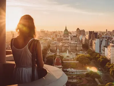 Eine Frau in einem weißen Trägerkleid steht mit dem Rücken zur Kamera und blickt über eine Stadt bei Sonnenuntergang. Im Vordergrund ist eine Stadtkulisse mit einem großen Gebäude mit grüner Kuppel und vielen anderen Gebäuden zu sehen, die in warmem orangefarbenem Licht gebadet sind.