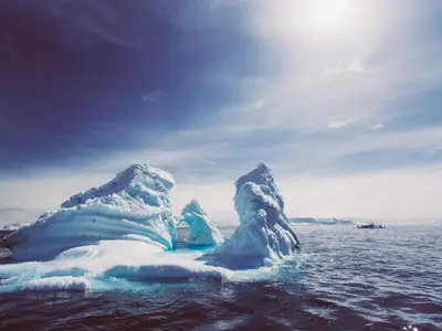 Mehrere massive Eisberge treiben in einem dunklen Meer, während die Celebrity Eclipse in der Ferne zu sehen ist. Der Himmel ist teils sonnig, teils bedeckt, über einer weiten, eisigen Landschaft.