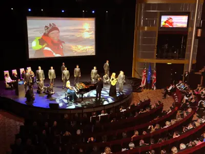 Festliche Bühnenshow auf der Celebrity Eclipse. Musiker und Sänger performen vor einer Leinwand mit einer Frau. Rote Publikumsreihen und Flaggen sind sichtbar.