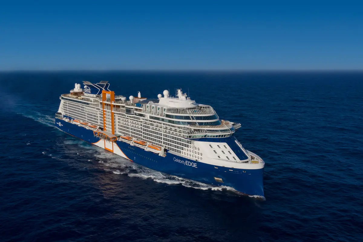 Luftaufnahme der Celebrity Edge auf offener See. Das Schiff mit blauem Rumpf und orangefarbenem Anbau gleitet durch das tiefblaue Meer.