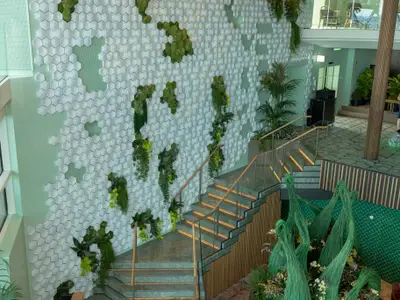 Modernes Interieur der Celebrity Edge. Eine Wand mit weißen Hexagon-Fliesen und Pflanzen dominiert den Raum. Eine beleuchtete Treppe führt zu einer unteren Ebene mit grünen Kunstwerken.