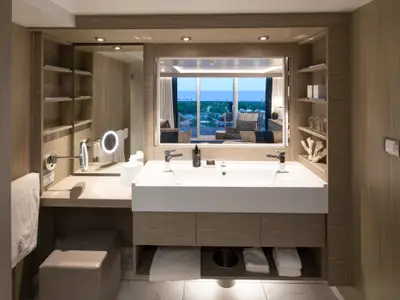 Ein elegantes Badezimmer mit Doppelwaschbecken und großem Spiegel. Im Spiegel ist ein Balkon mit Meerblick zu sehen. Das Badezimmer ist in hellen Holztönen gehalten und verfügt über offene Regale und eine Schminkkonsole.