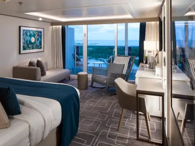 Innenansicht einer modernen Kreuzfahrtkabine auf der Celebrity Edge mit großem Bett, Sofa und Schreibtisch. Eine Glasfront führt zu einem Balkon mit Meer- und Küstenblick.