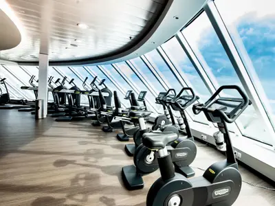 Modernes Fitnessstudio der Celebrity Edge mit Ergometern, Laufbändern und großen Fenstern zum Meer und Himmel. Sonnenlicht fällt auf den Holzboden.