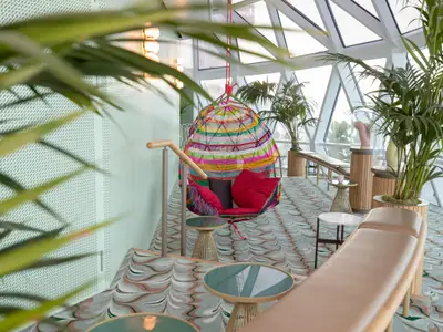 Eine helle, moderne Lounge auf der celebrity edge mit einem bunten Hängesessel, vielen Grünpflanzen in Holzgefäßen und großen Fenstern, die Meerblick bieten.