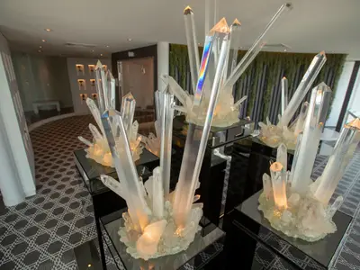 Eine Innenansicht einer modernen Lounge mit mehreren großen, beleuchteten Kristallinstallationen auf schwarzen Podesten. Im Hintergrund sind eine geschwungene Wand, eine weitere Sitzecke und eine grüne Wandverkleidung zu sehen. Der Boden hat ein geometrisches Muster.