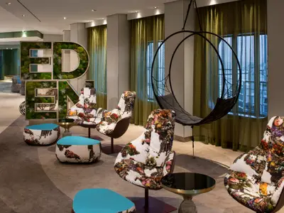 Eine Lounge mit vielen farbenfrohen Sesseln mit Kaktusmotiven und zwei passenden Hockern. Eine grosse Mooswand in Buchstabenform buchstabiert "EDEN". Ein schwarzer Hängesessel steht vor einem Fenster mit Gardinen.