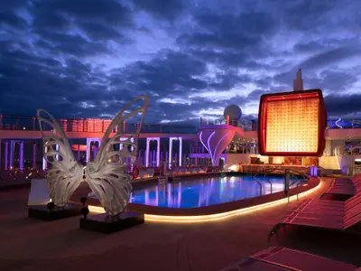 Abendliche Ansicht des Pooldecks der Celebrity Edge mit blau beleuchtetem Pool, eleganten Schmetterlingsflügel-Skulpturen und leuchtenden Installationen unter bewölktem Himmel.