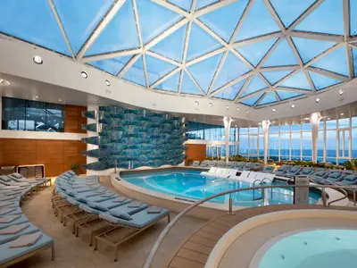 Das Solarium der Celebrity Edge: Ein großer Indoor-Poolbereich mit Liegen, Glasdach und Meerblick. Eine blaue Wellenwand ziert den Raum.