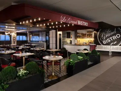 Innenaufnahme des Le Grand Bistro auf der Celebrity Edge mit eleganten Tischen und Stühlen. Eine Bäckereitheke, Pflanzkübel und Fenster mit Meerblick prägen den Raum.