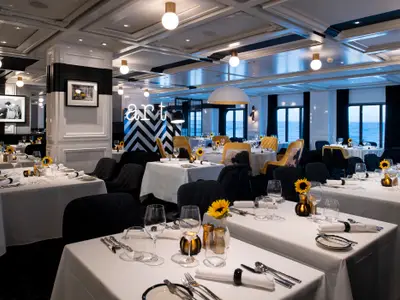 Modernes Restaurant auf der Celebrity Edge. Gedeckte Tische mit Sonnenblumen, schwarze und gelbe Stühle, schwarz-weiße Einrichtung. Große Fenster bieten Meerblick.