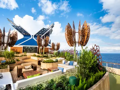 Blick auf den Dachgarten der Celebrity Edge mit Holzplattform, üppigen Pflanzen, modernen Lounge-Möbeln, Skulpturen und dem weiten Meer im Hintergrund unter blauem Himmel.