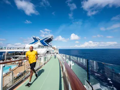 Ein Mann in gelbem Shirt joggt auf dem grünen Laufparcours an Deck der Celebrity Edge. Im Hintergrund das weite Meer, weitere Decks mit Pools und Passagiere unter blauem Himmel.