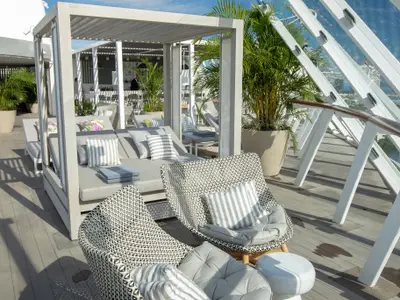 Sonniges Außendeck der Celebrity Edge mit modernen Lounge-Möbeln, Palmen und einem Barbereich. Im Hintergrund eine Glasfassade mit Küstenblick.
