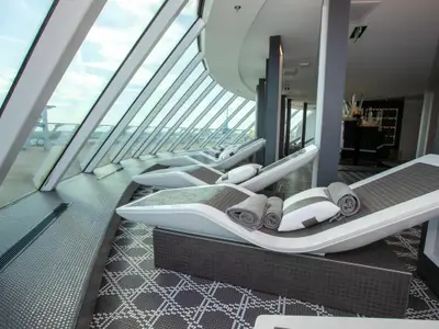 Innenansicht des eleganten Solariums der Celebrity Edge mit mehreren modernen, weißen Liegen. Große, schräge Fensterfronten geben den Blick auf den blauen Himmel frei.