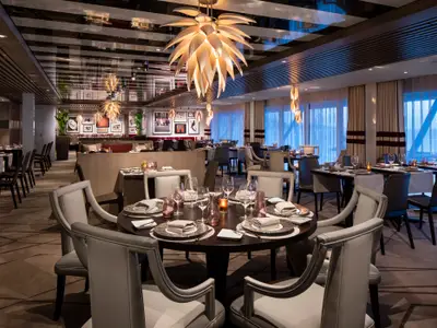Blick in das stilvolle Restaurant der Celebrity Edge. Elegant gedeckte Tische, helle Stühle, markante Deckenleuchte, große Fenster mit Ausblick prägen den modernen Raum. Warme Beleuchtung.