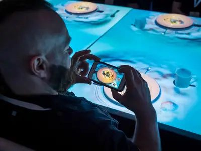 Mann filmt Essen in interaktivem Spezialitätenrestaurant mit blauer Lichtprojektion auf dem Tisch.