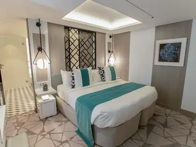 Ein modernes Schlafzimmer an Bord der Celebrity Edge. Es zeigt ein Doppelbett mit weißer Bettwäsche und einem türkisfarbenen Läufer. Stilvolle Wandleuchten, ein dekoratives Kopfteil und gemusterter Teppich prägen das Ambiente.