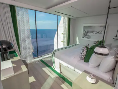 Helle Kabine der Celebrity Edge mit großem Panoramafenster zum Meer. Ein weißes Bett mit grünen Akzenten, Nachttische und eine moderne Lampe sind zu sehen. Die Einrichtung ist hell und geradlinig, mit einem grünen Streifen auf dem Boden.