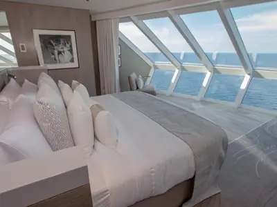 Moderne Kabine der Celebrity Edge: großes Bett mit vielen Kissen, dahinter Panorama-Fenster mit Blick auf das offene Meer. Helle Einrichtung und kleine Sitzecke.