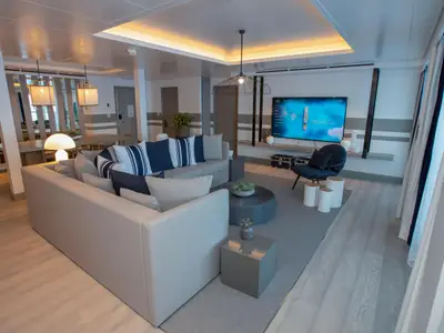 Heller, moderner Wohnbereich an Bord der Celebrity Edge mit großem L-förmigem Sofa, TV-Wand und eleganten Möbeln. Essbereich im Hintergrund.