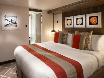 Moderne Kabine der Celebrity Edge. Großes Bett mit orange-beigem Läufer und Kissen. Drei gerahmte Blumenbilder über dem Bett. Heller Badzugang links. Eine weitere Wanddekoration ist sichtbar.