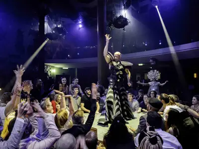 Bühnenshow auf der Celebrity Edge: Ein Darsteller in auffälligem Kostüm interagiert mit dem begeisterten Publikum. Violette Lichtstrahlen und andere Artisten sind sichtbar.
