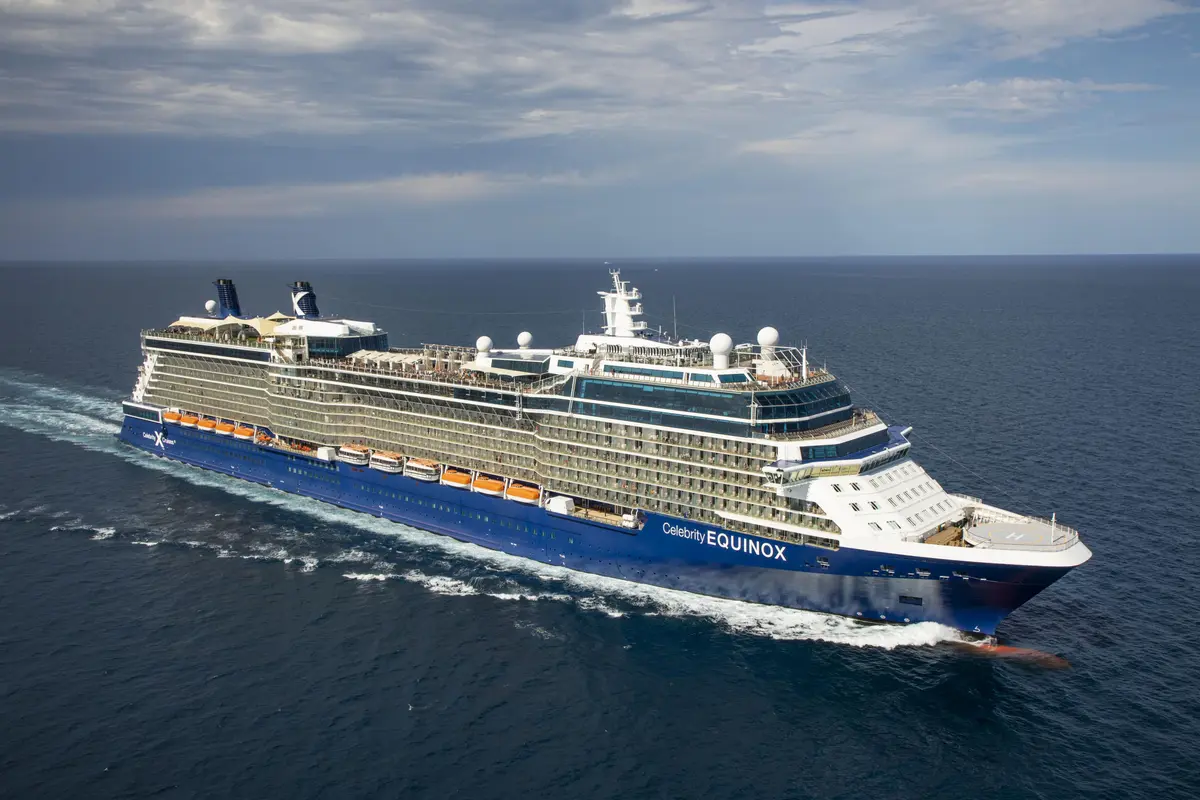 Ein blau-weißes Kreuzfahrtschiff namens Celebrity Equinox fährt schräg von links unten nach rechts oben auf dem tiefblauen Meer. Es hinterlässt eine weiße Gischtspur am Heck. Der Himmel ist teilweise bewölkt.