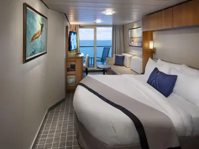 Innenansicht einer hellen Kabine auf dem Kreuzfahrtschiff Celebrity Equinox. Man sieht ein großes Bett, ein Sofa, einen Schreibtisch mit TV und einen Balkon mit Meerblick. Die Einrichtung ist in neutralen Farben mit blauen Akzenten und Holzelementen gehalten.