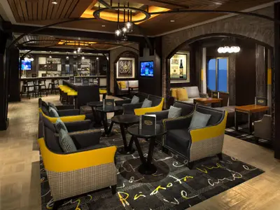 Blick in den Bar- und Lounge-Bereich der Celebrity Equinox mit einer langen Theke, gelb-schwarzen Sesseln, dunklem Holz und einem gemusterten Teppich. Blaue Fensterattrappen simulieren Meerblick.