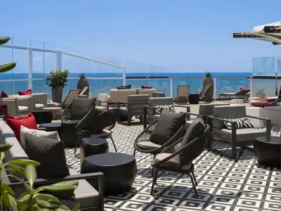 Modernes Sonnendeck mit grauen Lounge-Möbeln und roten Kissen auf schwarz-weiß gemustertem Boden. Meerblick durch Glasbrüstung unter blauem Himmel.