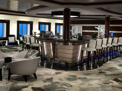 Blick in die stilvolle Bar und Lounge der Celebrity Equinox mit langer Theke, weißen Barhockern, bequemen Sesseln, gemustertem Teppich und großen Fenstern.