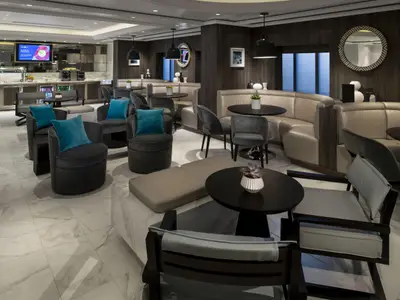 Die Lounge der Celebrity Equinox zeigt diverse Sitzgruppen mit Sesseln, Sofas und Tischen. Ein Buffetbereich ist im Hintergrund. Das moderne Design kombiniert dunkle Holztöne mit hellen Polstern und Fliesen.